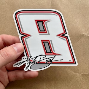 May include: A white sticker in the shape of the number 8, outlined in red, black, and gray. The number has a red interior with a gray rectangle. The sticker includes the signature "Kyle Busch" in black and the TM symbol.