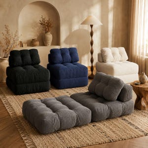 May include: A selection of corduroy modular seating in black, navy, cream, and grey. The image shows armchairs and ottomans on a woven rug. The furniture has a modern design, suitable for a living room. The seating is designed for comfort and style.