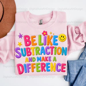 May include: A light pink crewneck sweatshirt featuring the colourful text BE LIKE SUBTRACTION AND MAKE A DIFFERENCE. The design includes a yellow smiley face, a pink flower, and small yellow and purple stars in a vibrant, multi-coloured font style.