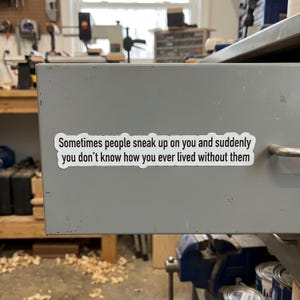 May include: A white sticker with black text is adhered to a gray metal drawer. The text reads, "Sometimes people sneak up on you and suddenly you don't know how you ever lived without them."