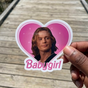 May include: A heart-shaped sticker featuring a portrait of a person with shoulder-length brown hair set against a bright pink background with white star accents. The word Babygirl is printed in white letters with a pink outline at the bottom.