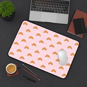 Peut inclure: Un tapis de bureau rectangulaire avec un motif répétitif de croissants dorés sur un fond rose clair parsemé de petits points blancs. Le tapis présente une bordure noire et est posé sur une surface sombre avec un ordinateur portable et un mug.