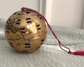 May include: A spherical brass incense burner featuring intricate cut-out patterns across its surface. The object is hinged at the center and includes a thin, braided dark pink cord with a matching tassel attached to the top for hanging purposes.