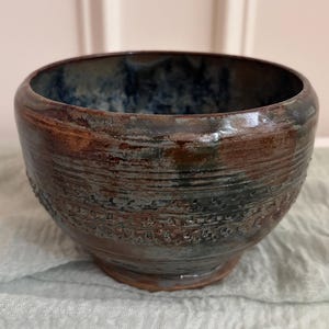 May include: A small, round ceramic bowl featuring a dark, mottled glaze with blended tones of deep brown, charcoal, and metallic blue. The exterior surface displays a textured, horizontal band of impressed markings around the center.