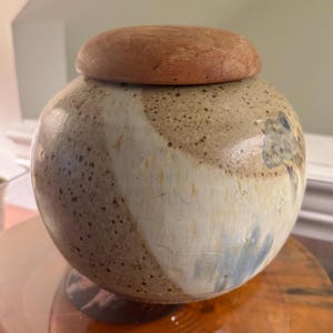 May include: A round ceramic jar with a wooden lid. The jar has a speckled, off-white and tan glaze with blue accents. The lid is a solid, reddish-brown color. The jar sits on a wooden base.