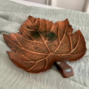 May include: A copper-colored metal dish shaped like a leaf with a textured surface and raised veins. The dish features a small stem-like handle and shows areas of dark oxidation and green patina throughout the metallic surface.