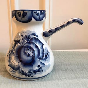 May include: A white ceramic coffee pot with a rounded body and a long, slender handle. The pot features hand-painted blue floral patterns and decorative scalloped blue trim around the top rim, showcasing a classic cobalt blue and white color scheme.