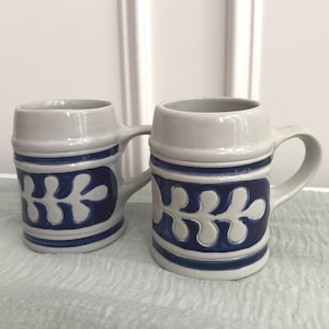 May include: Two light gray ceramic mugs with a glossy finish, each featuring a prominent dark blue band around the center. The blue band is decorated with a repeating white leaf-like pattern. Each mug has a sturdy handle and a smooth, rounded rim.