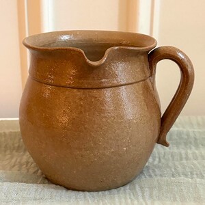 May include: A brown ceramic pitcher with a rounded body, a wide rim, and a curved handle. The surface has a textured, glossy finish with subtle variations in color. The pitcher features a small, integrated spout for pouring liquids.