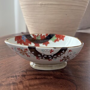 May include: A small, square-shaped ceramic bowl with a raised base. The bowl features a white interior with a decorative band of red, orange, and green leaves and flowers. The rim is trimmed with a dark brown band.
