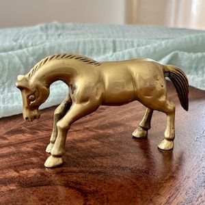 May include: A brass horse figurine in a trotting pose. The sculpture is a golden color with a dark tail and mane. The horse is standing on four legs, with its head lowered. The figurine is on a dark wooden surface.