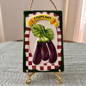 May include: A rectangular ceramic wall plaque featuring a painted illustration of three purple eggplants with green leaves. The background has a red and white checkered border, and a yellow banner at the top displays the word EGGPLANT in black.