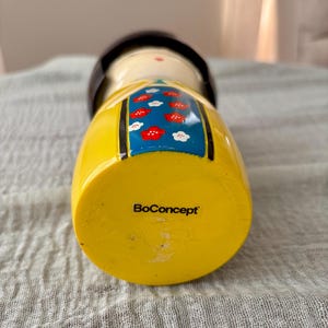 May include: A wooden kokeshi doll featuring a yellow base with the text BoConcept printed in black. The doll has a cream-colored face, a dark brown rounded top, and a blue central panel decorated with a pattern of small red and white flowers.