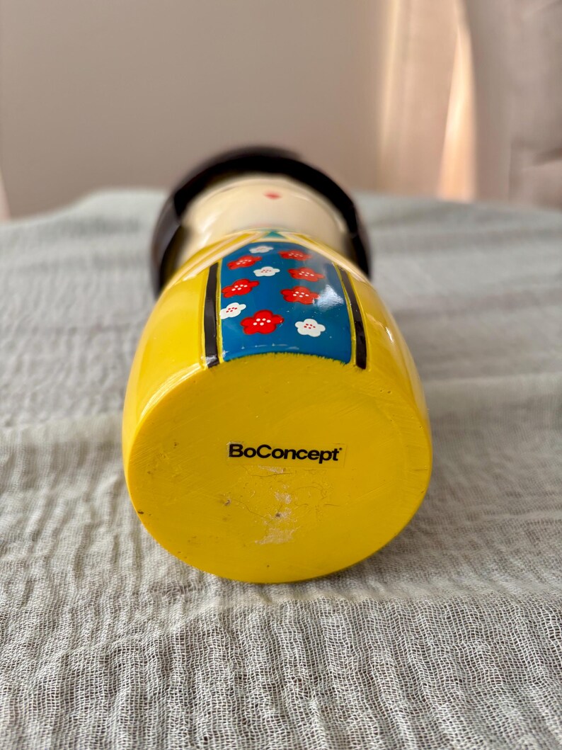 May include: A wooden kokeshi doll featuring a yellow base with the text BoConcept printed in black. The doll has a cream-colored face, a dark brown rounded top, and a blue central panel decorated with a pattern of small red and white flowers.