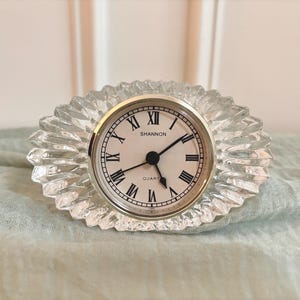 May include: An oval-shaped Shannon quartz clock featuring a clear, faceted glass frame with a sunburst pattern. The clock face is white with black Roman numerals and thin black hands, set within a polished silver-toned metal circular bezel.