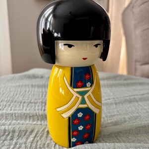 May include: A Kokeshi doll featuring a smooth, glossy black bob hairstyle and a pale face with simple facial features. The body is painted in a bright yellow with a central blue panel decorated with small red and white floral patterns.