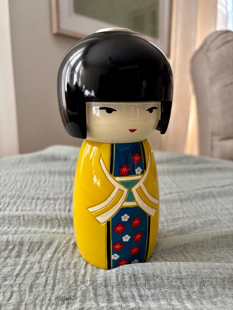 May include: A Kokeshi doll featuring a smooth, glossy black bob hairstyle and a pale face with simple facial features. The body is painted in a bright yellow with a central blue panel decorated with small red and white floral patterns.