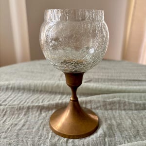 May include: A clear glass candle holder with a crackled texture, resting on a bronze-colored metal base. The base has a flared, circular foot and a slender stem. The candle holder is designed to hold a small candle.