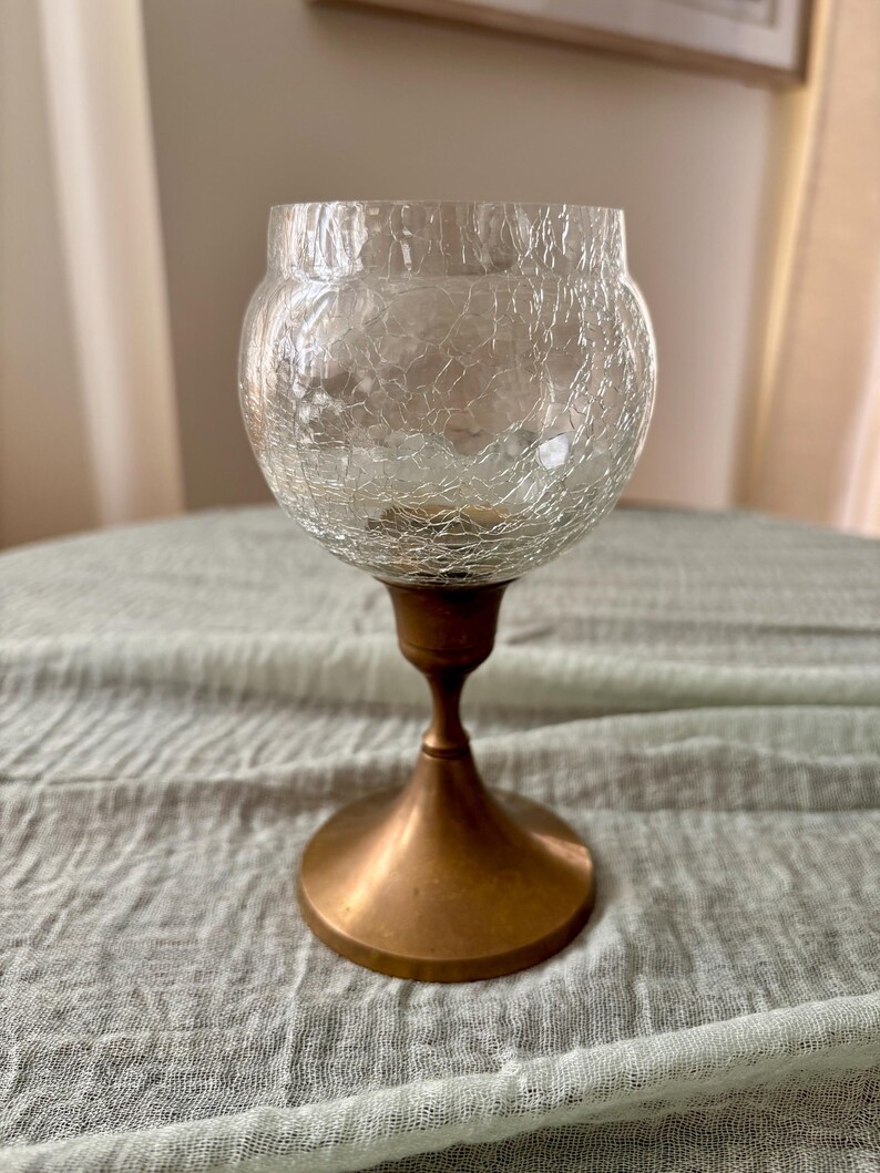May include: A clear glass candle holder with a crackled texture, resting on a bronze-colored metal base. The base has a flared, circular foot and a slender stem. The candle holder is designed to hold a small candle.