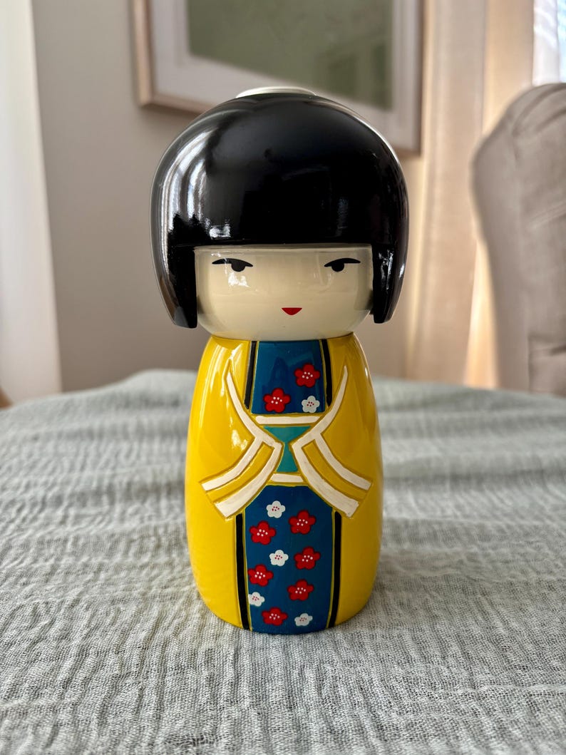 May include: A Kokeshi-style doll with a glossy black bob hairstyle and a smooth, pale face. The body is painted bright yellow with a central blue panel featuring a repeating pattern of small red and white flowers and white trim details.