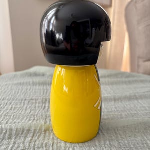 May include: A Kokeshi-style doll featuring a smooth, glossy black rounded head and a bright yellow cylindrical body. The figure has a minimalist design with a white and grey graphic detail on the side of the yellow torso, showcasing a clean, polished finish.