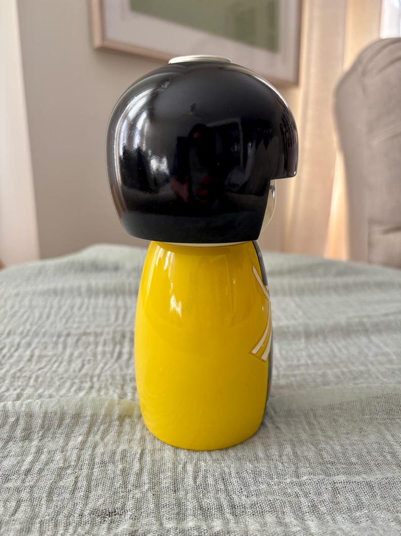 May include: A Kokeshi-style doll featuring a smooth, glossy black rounded head and a bright yellow cylindrical body. The figure has a minimalist design with a white and grey graphic detail on the side of the yellow torso, showcasing a clean, polished finish.