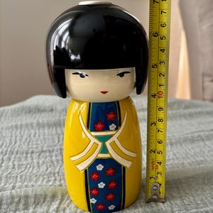 May include: A Kokeshi doll with a glossy black bob hairstyle, a yellow kimono featuring a blue panel with red and white floral patterns, and a measuring tape showing a height of approximately 8.5 inches from the base to the top of the head.
