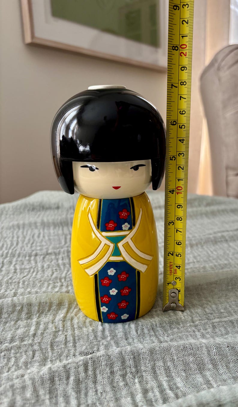 May include: A Kokeshi doll with a glossy black bob hairstyle, a yellow kimono featuring a blue panel with red and white floral patterns, and a measuring tape showing a height of approximately 8.5 inches from the base to the top of the head.