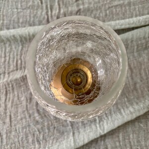 May include: A clear, crackled glass votive candle holder with a gold-colored metal base. The candle holder has a round shape and a frosted rim. The glass has a textured, web-like pattern.