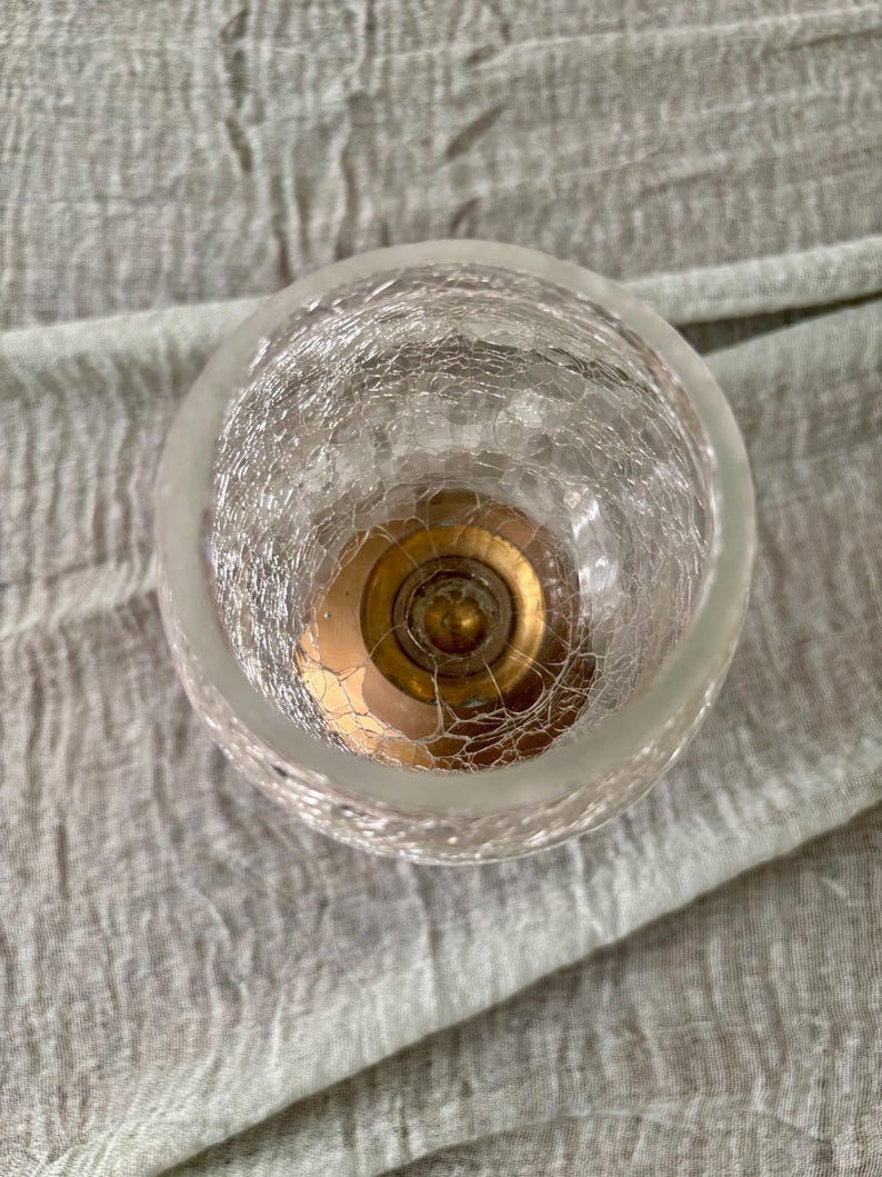 May include: A clear, crackled glass votive candle holder with a gold-colored metal base. The candle holder has a round shape and a frosted rim. The glass has a textured, web-like pattern.