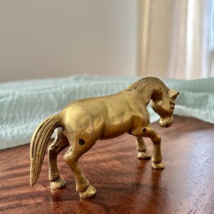 May include: A small, antique brass horse figurine. The horse is posed in a walking stance, with its head slightly lowered. The figurine has a detailed mane and tail, and is set on a dark wooden surface.