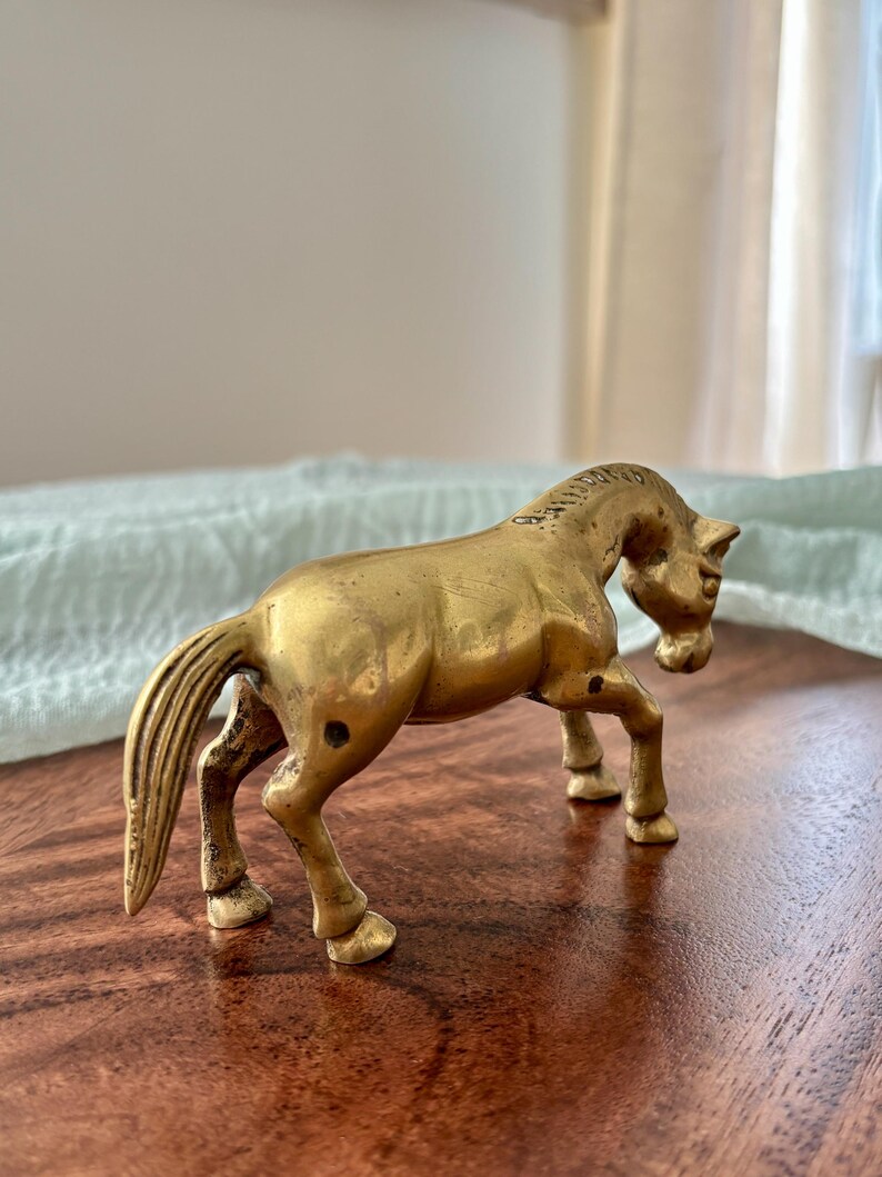 May include: A small, antique brass horse figurine. The horse is posed in a walking stance, with its head slightly lowered. The figurine has a detailed mane and tail, and is set on a dark wooden surface.