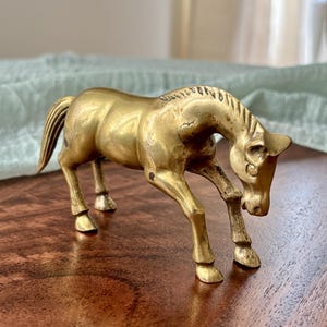 May include: A vintage brass horse figurine in a bowing pose. The sculpture has a detailed mane and tail, with its head lowered towards the ground. The horse is a warm gold color and stands on four legs.