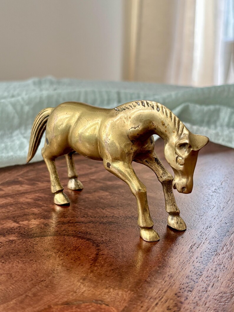 May include: A vintage brass horse figurine in a bowing pose. The sculpture has a detailed mane and tail, with its head lowered towards the ground. The horse is a warm gold color and stands on four legs.