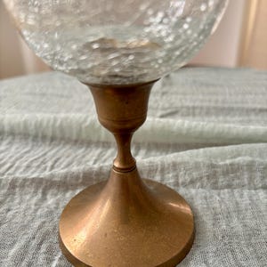 May include: A clear glass globe with a textured surface sits atop a bronze-colored metal base. The base has a flared, circular foot and a slender stem, creating a vintage aesthetic. The glass globe appears to be a decorative item.