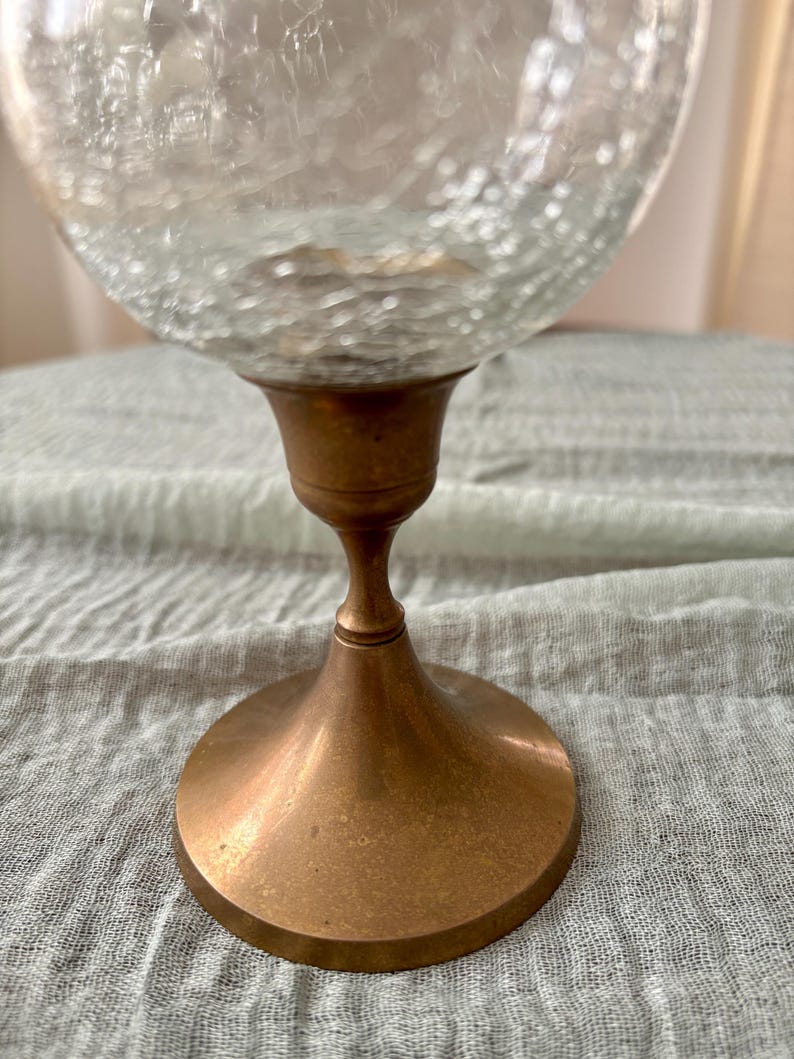 May include: A clear glass globe with a textured surface sits atop a bronze-colored metal base. The base has a flared, circular foot and a slender stem, creating a vintage aesthetic. The glass globe appears to be a decorative item.
