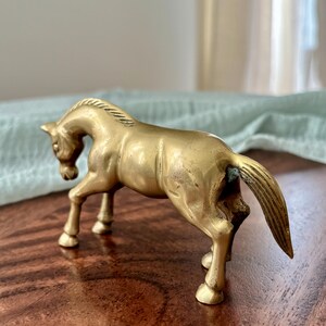 May include: A brass horse figurine in a standing pose. The sculpture is a golden color and has detailed features, including a flowing mane and tail. The horse is on a wooden surface.