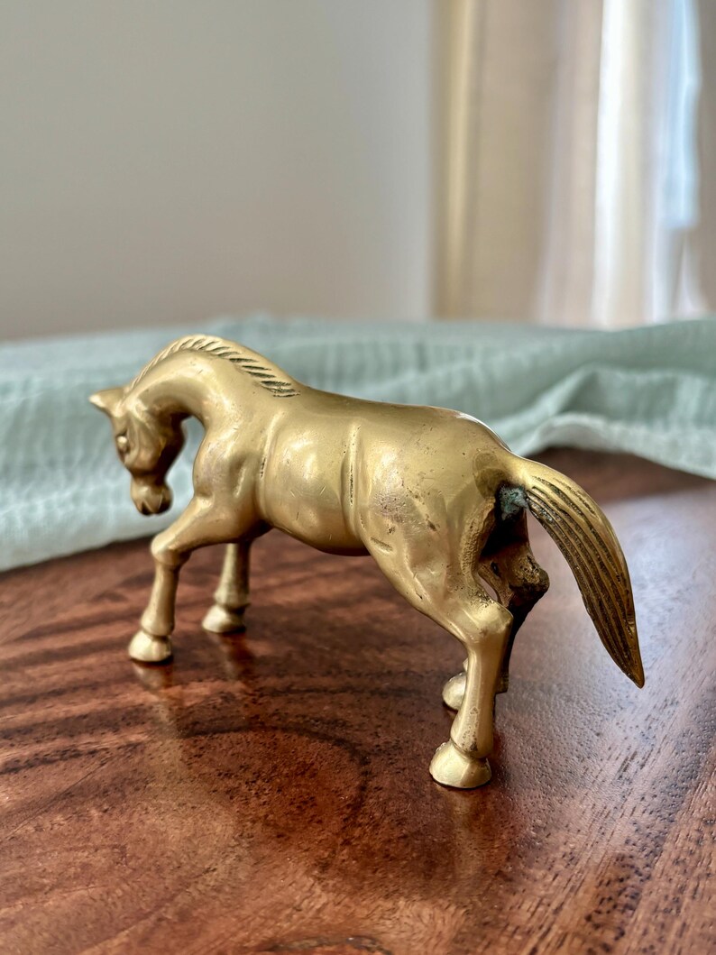 May include: A brass horse figurine in a standing pose. The sculpture is a golden color and has detailed features, including a flowing mane and tail. The horse is on a wooden surface.