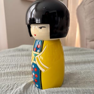May include: A Kokeshi doll featuring a smooth, black, rounded bob hairstyle and a cream-colored face with simple painted features. The body is a bright yellow cylinder decorated with a blue panel displaying a pattern of small red and white flowers.