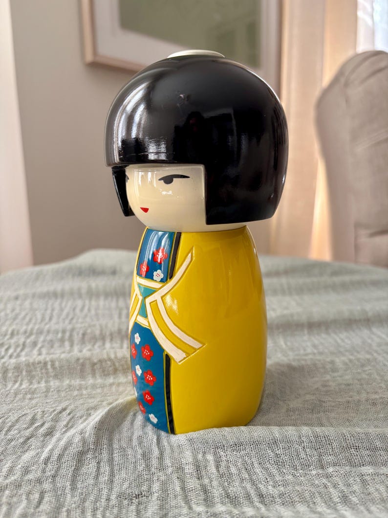 May include: A Kokeshi doll featuring a smooth, black, rounded bob hairstyle and a cream-colored face with simple painted features. The body is a bright yellow cylinder decorated with a blue panel displaying a pattern of small red and white flowers.