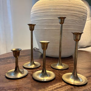 May include: Five brass candle holders of varying heights are arranged on a wooden surface. Each holder has a circular base and a slender stem leading to a cup-shaped top. A large, textured white lamp base is in the background.