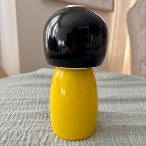May include: A smooth, rounded wooden figurine featuring a glossy black top section that resembles a stylized hairstyle and a solid, bright yellow base. The object has a cylindrical shape with a slightly wider bottom, showcasing a clean, polished finish.
