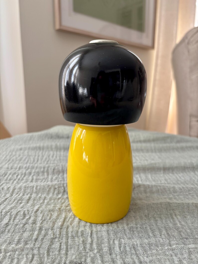 May include: A smooth, rounded wooden figurine featuring a glossy black top section that resembles a stylized hairstyle and a solid, bright yellow base. The object has a cylindrical shape with a slightly wider bottom, showcasing a clean, polished finish.