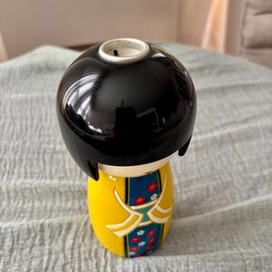 May include: A ceramic kokeshi doll candle holder with a glossy black bob hairstyle. The body is bright yellow with a central blue panel featuring a red and white floral pattern. The top of the head has a circular opening for a candle insert.