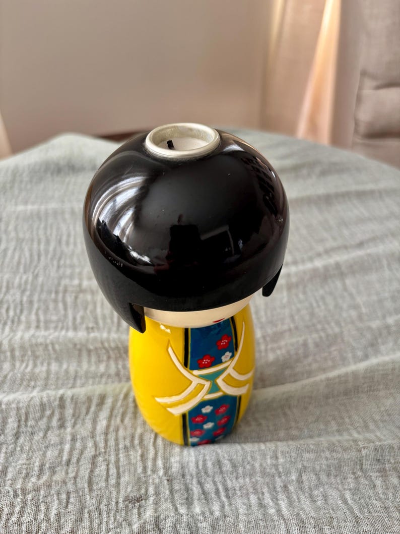 May include: A ceramic kokeshi doll candle holder with a glossy black bob hairstyle. The body is bright yellow with a central blue panel featuring a red and white floral pattern. The top of the head has a circular opening for a candle insert.