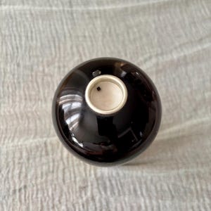May include: A spherical, dark brown ceramic vessel with a smooth, glossy finish. The top features a circular opening with a light-colored interior rim and a small, dark square detail visible inside the center of the opening.