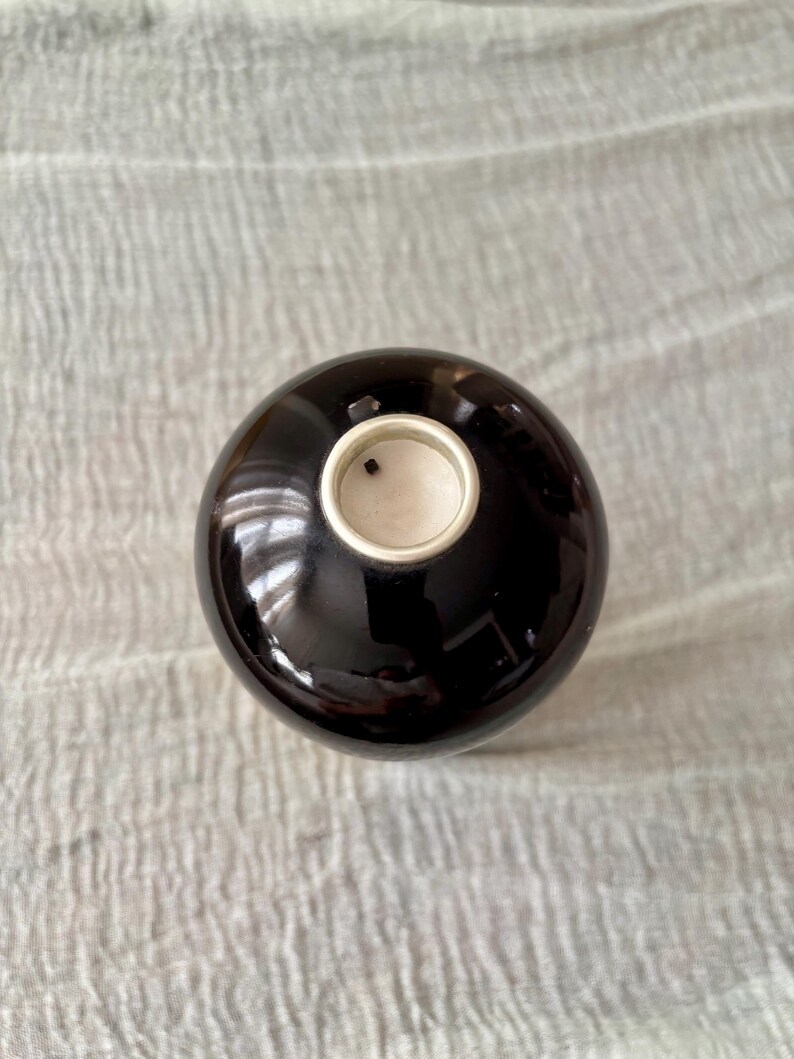 May include: A spherical, dark brown ceramic vessel with a smooth, glossy finish. The top features a circular opening with a light-colored interior rim and a small, dark square detail visible inside the center of the opening.