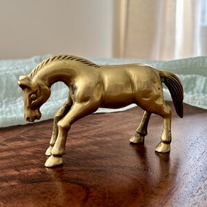 May include: A vintage brass horse figurine. The sculpture depicts a horse in a trotting pose, with its head lowered. The horse is a warm gold color, with a detailed mane and tail. This decorative item is suitable for home decor.