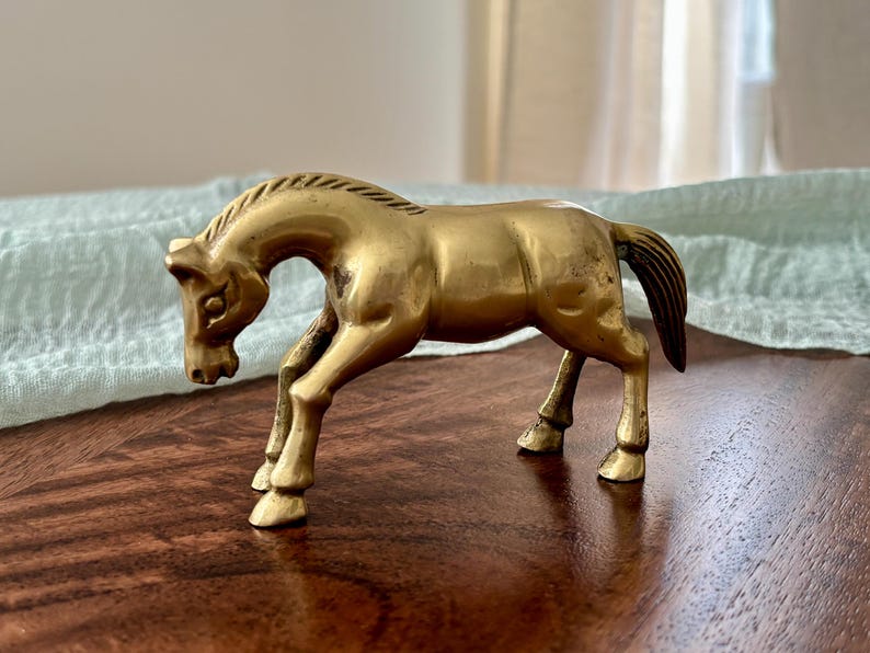 May include: A vintage brass horse figurine. The sculpture depicts a horse in a trotting pose, with its head lowered. The horse is a warm gold color, with a detailed mane and tail. This decorative item is suitable for home decor.