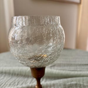 May include: A clear glass candle holder with a crackled texture, resting on a bronze-colored metal base. The candle holder has a rounded bowl shape and a slender stem, creating an elegant aesthetic.
